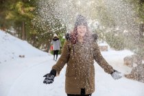 Веселая женщина в верхней одежде, стоящая в зимнем лесу и выбрасывающая снег, веселясь — стоковое фото