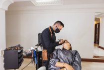 Homme masqué enduisant fond de teint de femme blonde pendant le travail dans un studio de maquillage professionnel — Photo de stock