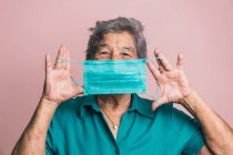 Lächelnde ältere Frauen, die ihren Mund mit blauer medizinischer Schutzmaske vor Coronavirus bedecken, während sie im Studio auf rosa Hintergrund in die Kamera schauen — Stockfoto