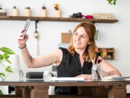 Веселая женщина-дизайнер сидит за столом с художественными принадлежностями и делает самоснимок на смартфоне в рабочем пространстве — стоковое фото