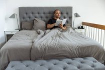 Pensivo macho sentado em cama macia de manhã e lendo história interessante no livro após o despertar — Fotografia de Stock
