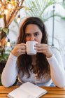 Молодая длинноволосая латиноамериканка наслаждается вкусным ароматным кофе из керамической чашки, отдыхая в уютном кафе с зелеными растениями — стоковое фото