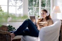 Inhalt weiblich im drahtlosen Headset surft im Internet auf dem Tablet, während sie zu Hause im Sessel Lieder hört — Stockfoto