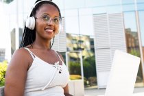 Donna afroamericana positiva in cuffia seduta con laptop in strada a Barcellona e che si gode le canzoni guardando altrove — Foto stock