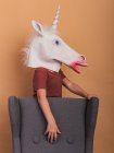 Side view of anonymous kid in decorative unicorn mask with open mouth touching armchair on beige background — Stock Photo