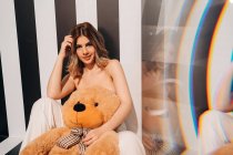 Fröhliche barfüßige Frau in weißen Hosen mit weichem Bären sitzt auf gestreiftem Boden und blickt in die Kamera — Stockfoto