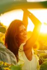 Щаслива молода іспанка в стильній жовтій сукні стоїть з піднятими руками серед квітучих соняшників на сільському полі в сонячний літній день дивлячись на камеру — стокове фото