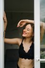Содержание стройная женщина в черном белье стоит возле стеклянной двери, ведущей на балкон и глядя в сторону — стоковое фото