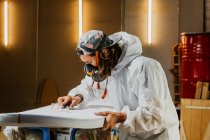 Side view of male shaper in safety costume and respirator polishing surface of surf board with hand plane in workshop — Stock Photo