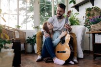 Content female Cheerful tattooed male musician playing guitar near Cheerful tattooed male musician while looking at each other in armchair in house room — Stock Photo