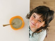 Dall'alto di bambino allegro che si siede a tavola con boccia di minestra alla panna e guarda la macchina fotografica durante pranzo in cucina a casa — Foto stock