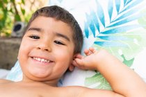 Милий маленький хлопчик посміхається і дивиться на камеру, лежачи на барвистому ліжку в літній день — стокове фото