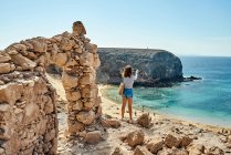 Vista trasera de una turista irreconocible con ropa casual parada cerca de ruinas de piedra y disparando mar turquesa en el día de verano en Fuerteventura, España - foto de stock