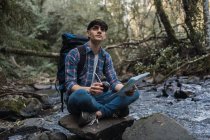 Viaggio zaino in spalla maschile con compagno di bere e carta mappa seduta sulla roccia vicino al fiume nei boschi e guardando in alto — Foto stock