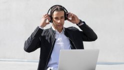 Young African American male manager with braids in formal suit and wireless headphones looking at camera while working on laptop on urban street — Stock Photo