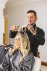 Male hairstylist using iron to curl blond locks of female customer during work in beauty salon — Stock Photo