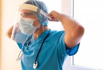 Чоловік лікар одягає захисний пластиковий щит обличчя під час роботи в клініці і дивиться вбік — стокове фото