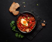 Chakchouka von oben mit sonnigem Ei in köstlicher Tomatensauce mit Roggenbrot in Schüssel zwischen frischem Koriander auf dunklem Hintergrund — Stockfoto