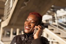 Vista lateral da bela mulher negra afro conversando com seu smartphone enquanto sorri com fundo embaçado em um dia ensolarado — Fotografia de Stock