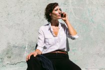 Full body of delighted female in white formal shirt and pants sitting on concrete border and having video call — Stock Photo
