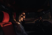 Боковой вид любящей пары в солнцезащитных очках, обнимающейся в винтажном автомобиле во время прогулки на природе вечером — стоковое фото