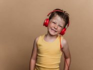 Zufriedener Frühchen-Junge mit roten Kopfhörern hört Musik auf braunem Hintergrund im Studio — Stockfoto