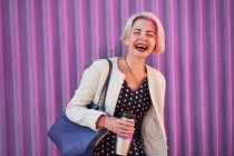 Позитивна неформальна жінка з пофарбованим коротким волоссям, що стоїть з напоєм в екологічно чистій чашці на фоні фіолетової стіни в місті і закритих очей — стокове фото