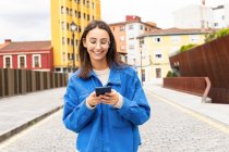 Усміхнена жінка зі спантеличеним волоссям, що йде по вулиці міста в вітряний день і використовує смартфон — стокове фото