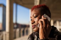 Vista lateral de una hermosa mujer afro negra hablando con su teléfono inteligente mientras mira hacia otro lado en un fondo borroso en un día soleado - foto de stock