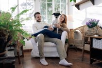 Bearded man with smiling girlfriend embracing purebred dog while resting in armchair against window in house room — Stock Photo