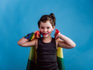 Fille souriante avec joue peinte levant les bras avec drapeau multicolore sur fond bleu vif — Photo de stock