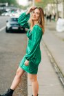 Femme insouciante en robe verte tendance debout avec un bras levé et touchant la tête dans la rue et regardant la caméra — Photo de stock