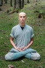 Bald man in traditional clothes sitting on grass in Lotus pose and meditating during kung fu training in forest — Stock Photo