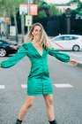 Unbekümmerte Frau im trendigen grünen Kleid steht mit ausgestreckten Armen auf der Straße und blickt in die Kamera — Stockfoto