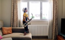 Seitenansicht eines barfüßigen Kindes mit geschlossenen Augen, das Saxofon spielt, während es tagsüber zu Hause auf der Couch steht — Stockfoto
