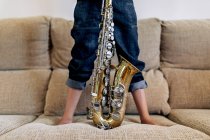 Cropped anonymous thoughtful barefoot child in hat with saxophone standing on sofa against window at home — Stock Photo