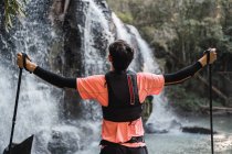 Rückansicht eines männlichen Wanderers mit Wanderstöcken in ausgestreckten Händen, der mit geschlossenen Augen in der Nähe eines Wasserfalls im Wald steht und die Freiheit genießt — Stockfoto