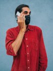 Hombre étnico en calma máscara protectora hablando en el teléfono móvil, mientras que de pie contra la pared azul en la ciudad durante coronavirus - foto de stock
