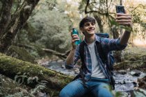 Durstige männliche Wanderer mit Rucksack trinken Wasser aus der Flasche, während sie Selfie auf einem Felsen in der Nähe des Wasserfalls im Wald sitzen und in der Pause wegschauen — Stockfoto