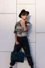 Jeune homme vaniteux en tenue élégante avec sac à main de dame debout sur le mur carrelé tout en regardant loin — Photo de stock