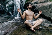 Friedliches Männchen ohne Hemd sitzt in Padmasana mit Mudra-Händen und geschlossenen Augen, während es Yoga macht und auf nassen Felsen in der Nähe des Wasserfalls meditiert — Stockfoto