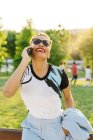 Cheerful African American female with red lips in trendy wear talking on cellphone while looking up in park — Stock Photo