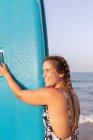 Seitenansicht einer nassen, glücklichen Surferin, die im Sommer mit blauem SUP-Board am Sandstrand steht und wegschaut — Stockfoto