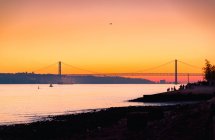 Terraplén abarrotado situado cerca del río Tajo con ferry cerca de la silueta del puente 25 de Abril contra el cielo naranja al atardecer en Lisboa, Portugal - foto de stock