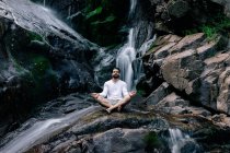 Pacifico macho sentado em Padmasana com mãos de mudra e olhos fechados enquanto faz ioga e meditar na rocha molhada perto de cachoeira — Fotografia de Stock
