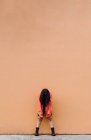 Cuerpo completo de joven mujer anónima cubriendo la cara con el pelo largo y castaño inclinándose hacia adelante mientras está de pie contra la pared naranja - foto de stock