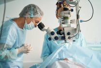 Взрослая женщина-врач в стерильной маске и декоративной медицинской шапке смотрит через хирургический микроскоп на коллегу по урожаю в больнице — стоковое фото