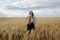 Позитивная молодая женщина в белой рубашке и красном галстуке, стоящая с руками позади пшеничных шипов в сельской местности — стоковое фото