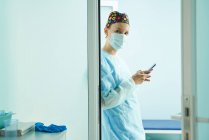 Побочный обзор женщины-врача в медицинской форме и стерильной маске текстовых сообщений на мобильный телефон в клинике — стоковое фото