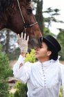 Grave adulto nero femmina in cappello ed elegante vestito con guanti petting cavallo marrone con gli occhi chiusi vicino a piante verdi e alberi in campagna — Foto stock